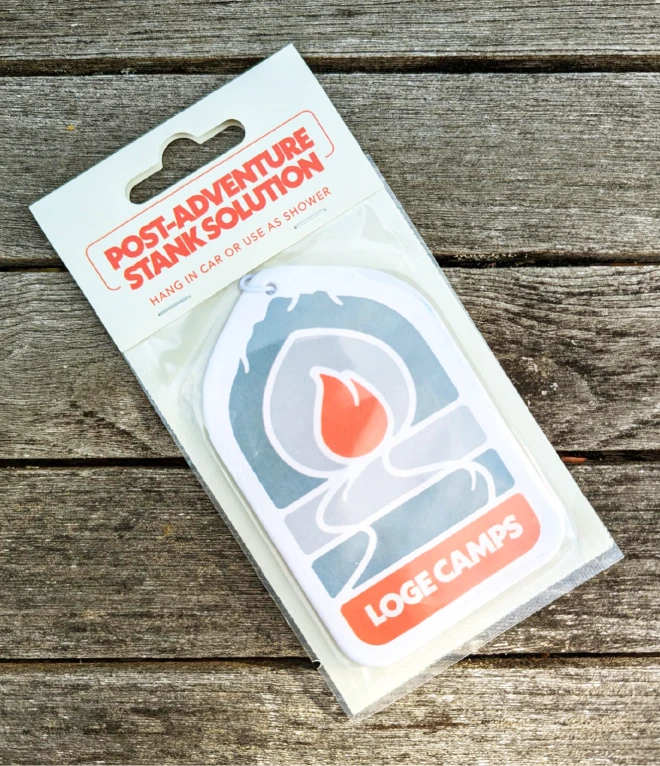 A packaged LOGE Camps air freshener in the shape of a stylized campfire with the label "Post-Adventure Stank Solution." The entire package is placed on a rustic wooden surface.