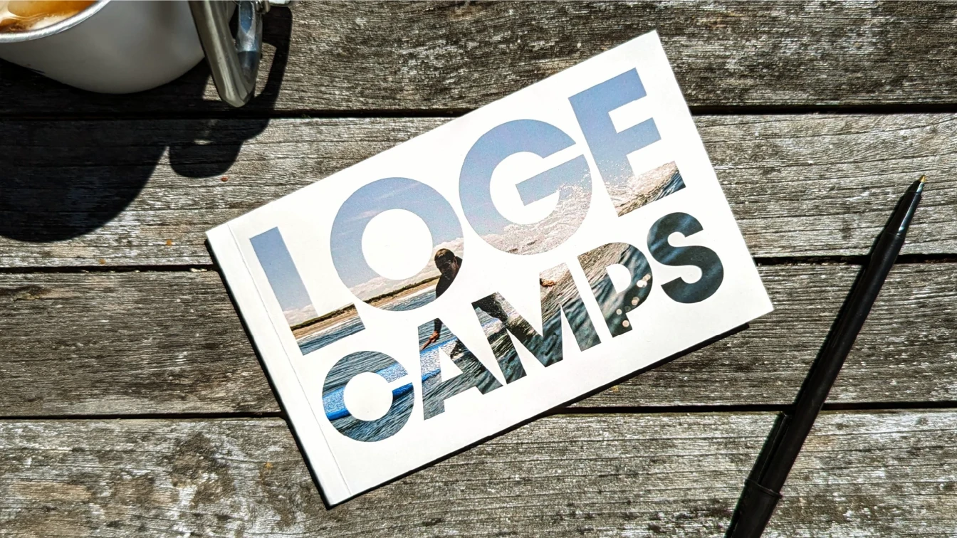A postcard with text "LOGE Camps" cut out, revealing a scenic beach view through the cutouts. The postcard is on a wooden surface next to a mug and a black pen.