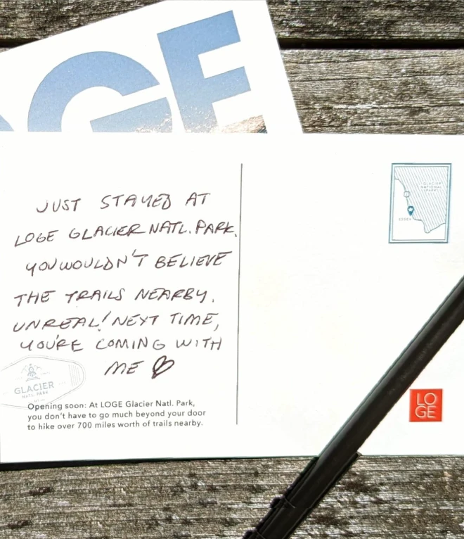 A postcard on a rustic wooden surface with a handwritten message. The message and postcard printed text mention the LOGE Glacier National Park. LOGE logo is on the bottom right.