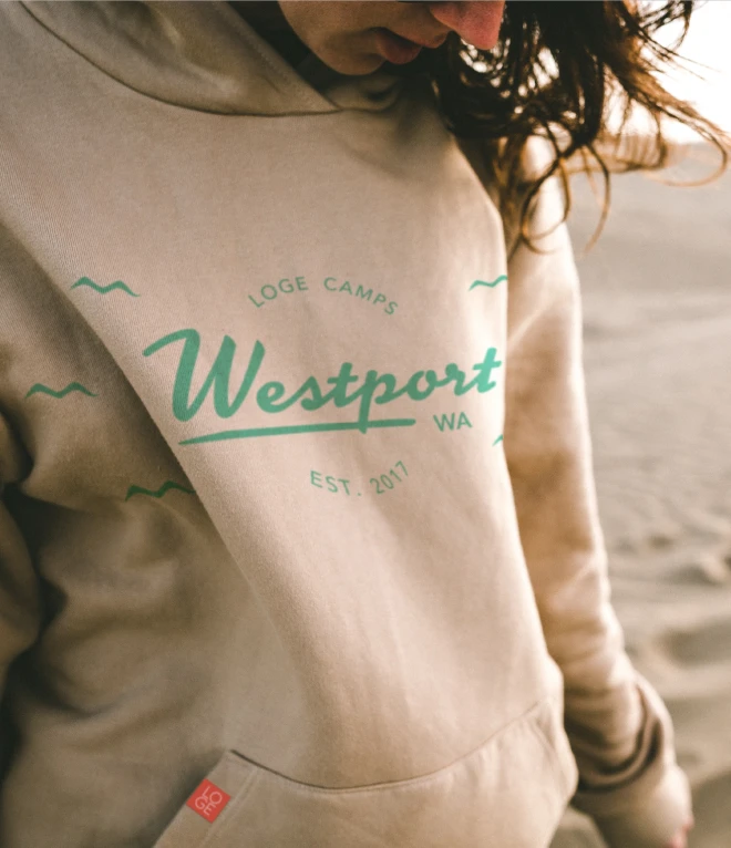 A person wearing a beige hoodie outdoors in sand with the LOGE Camps logo and the text "Westport, WA" printed on the front in green. The hoodie also includes "Est. 2017" under the Westport text.