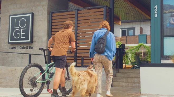 Four people ride bikes on a gravel path between cabins with blue metal roofs, surrounded by green trees