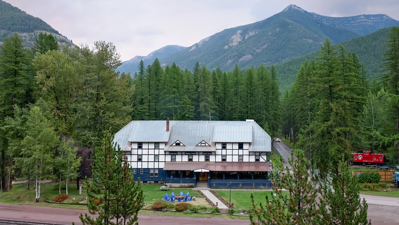 Rustic lodge in a pine forest with mountainous terrain behind, featuring a grassy yard and outdoor seating area