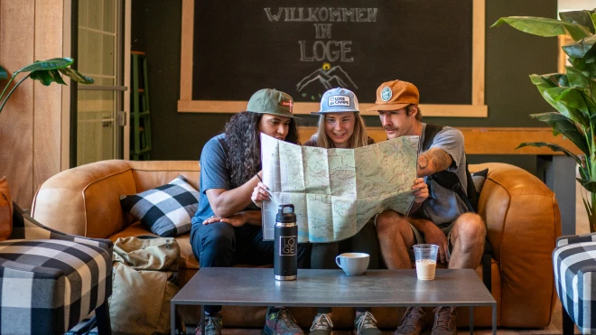 Three friends sitting on a couch studying a map with drinks on the table, under a sign reading Willkommen in LOGE