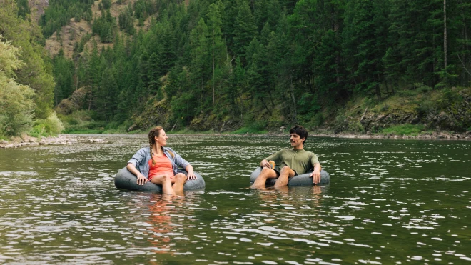 Two people relaxing on inner tubes in a calm river surrounded by forested hills and trees