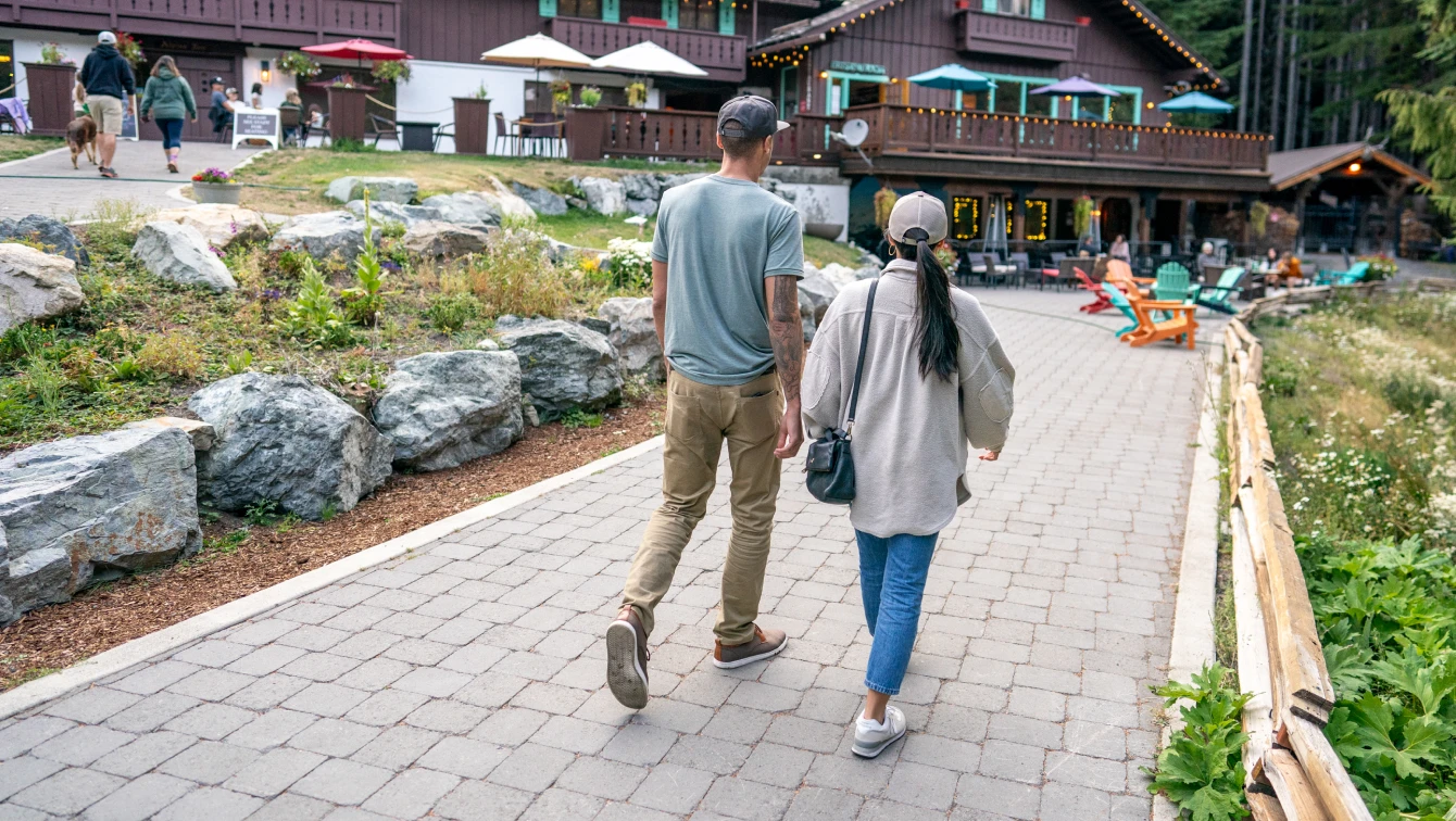 Couple walking on a stone path toward a lodge with patio seating, umbrellas, and people relaxing outside
