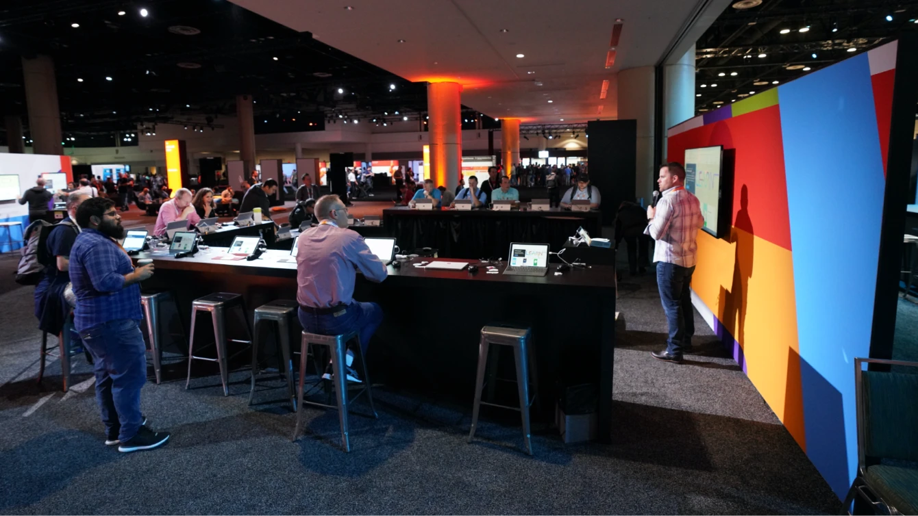 A lively scene at a Microsoft Learn event with attendees seated at a long counter, engaging with laptops and tablets and a speaker stands at the front.