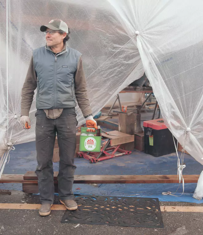 Man in workwear holding a Redhook IPA six-pack outside a workshop with plastic sheeting in the background.