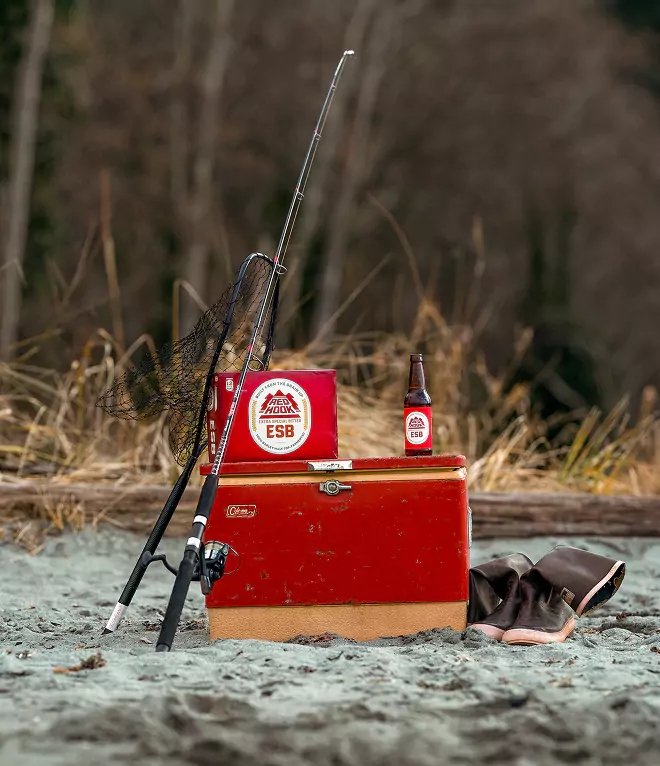 Fishing gear, Redhook ESB beer, and boots set on a red cooler at a sandy riverside spot with dry grass in the background.