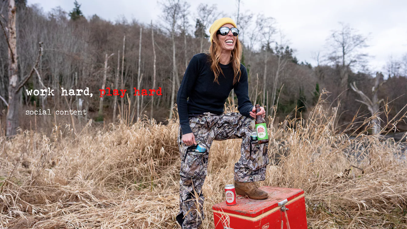 Smiling man outdoors holding a Redhook IPA bottle and a beer can, with text "work hard, play hard" overlayed.