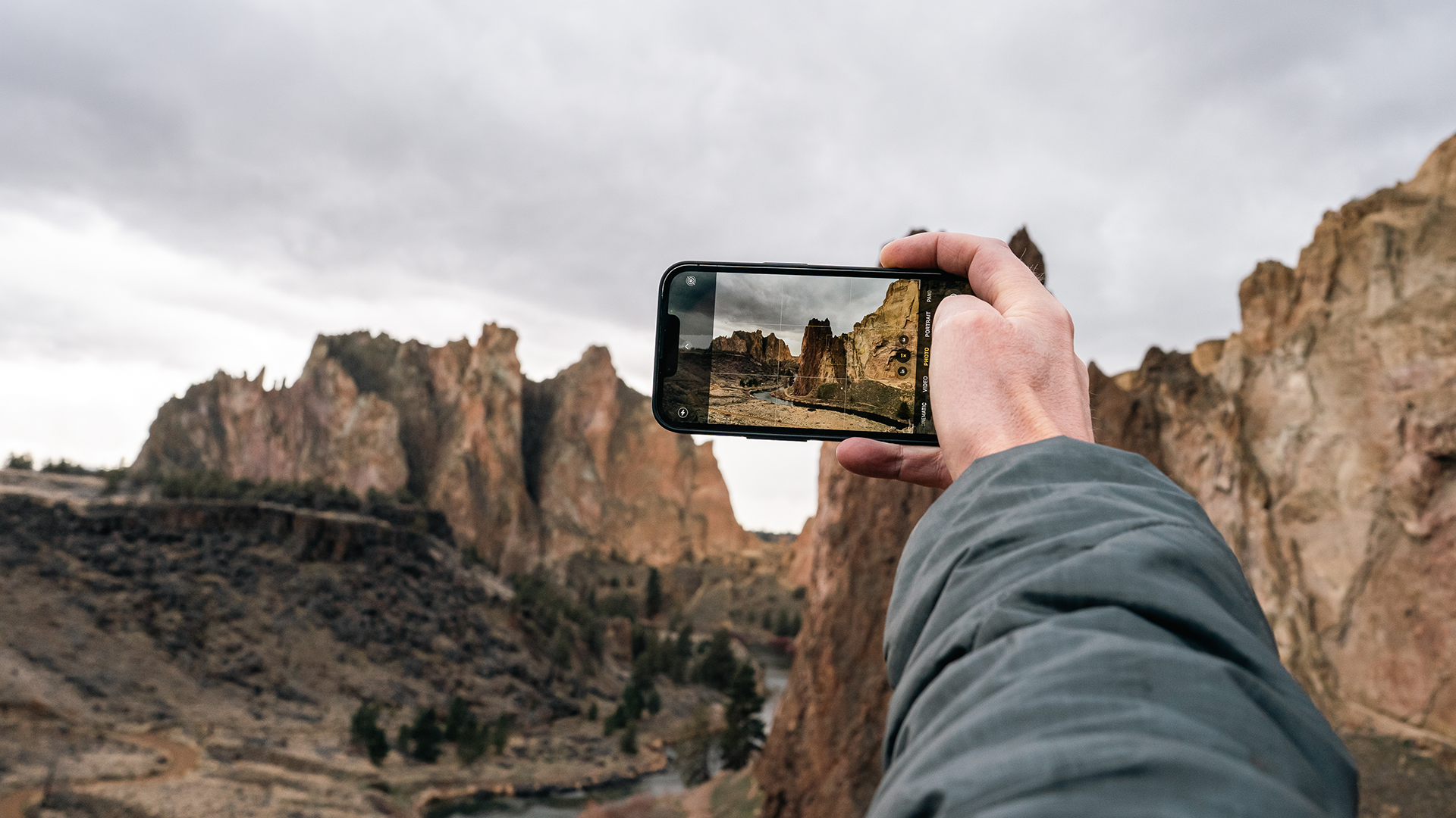 A man takes a photo using a mobile phone of a canyon with a river running through it.