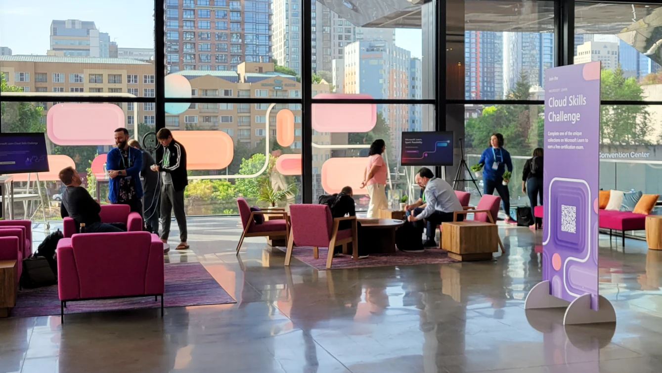 A bright and modern convention center lounge area with large floor-to-ceiling windows showcasing an urban skyline. A sign in the foreground promotes the "Cloud Skills Challenge".