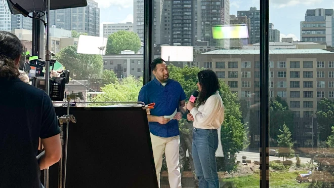 A live broadcast featuring two presenters standing in front of large windows with an urban skyline behind them and a camera and crew in the foreground.