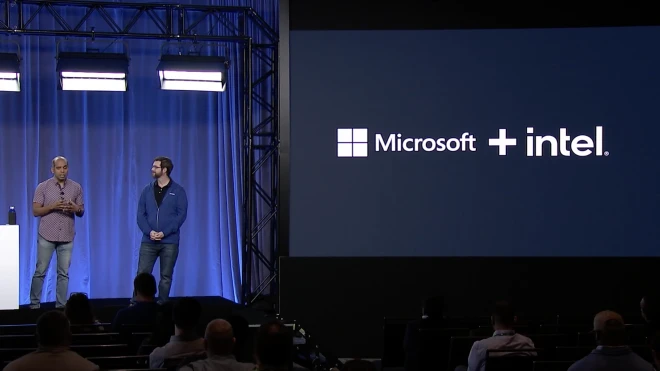 Two speakers present at a conference on stage next to a backdrop screen displaying the logos for a partnership between Microsoft and Intel.