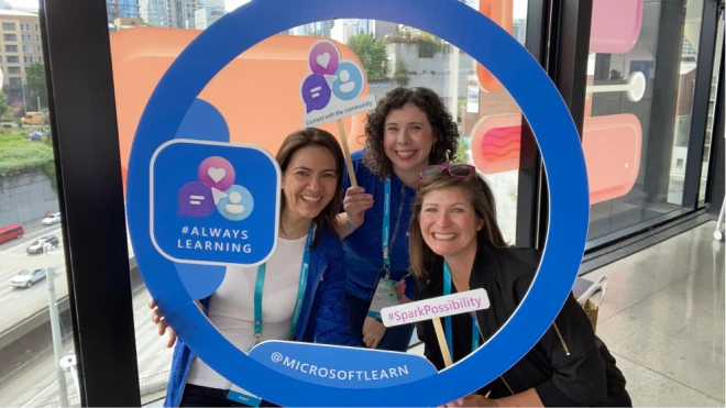 Three smiling women pose inside a large, circular blue frame that reads "@MICROSOFTLEARN". The background shows large windows with colorful, abstract decals and an urban cityscape.