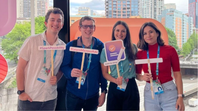 Four smiling individuals pose together, holding signs with messages in front of large windows with a cityscape in the background. The signs focus on Microsoft, AI, and learning.